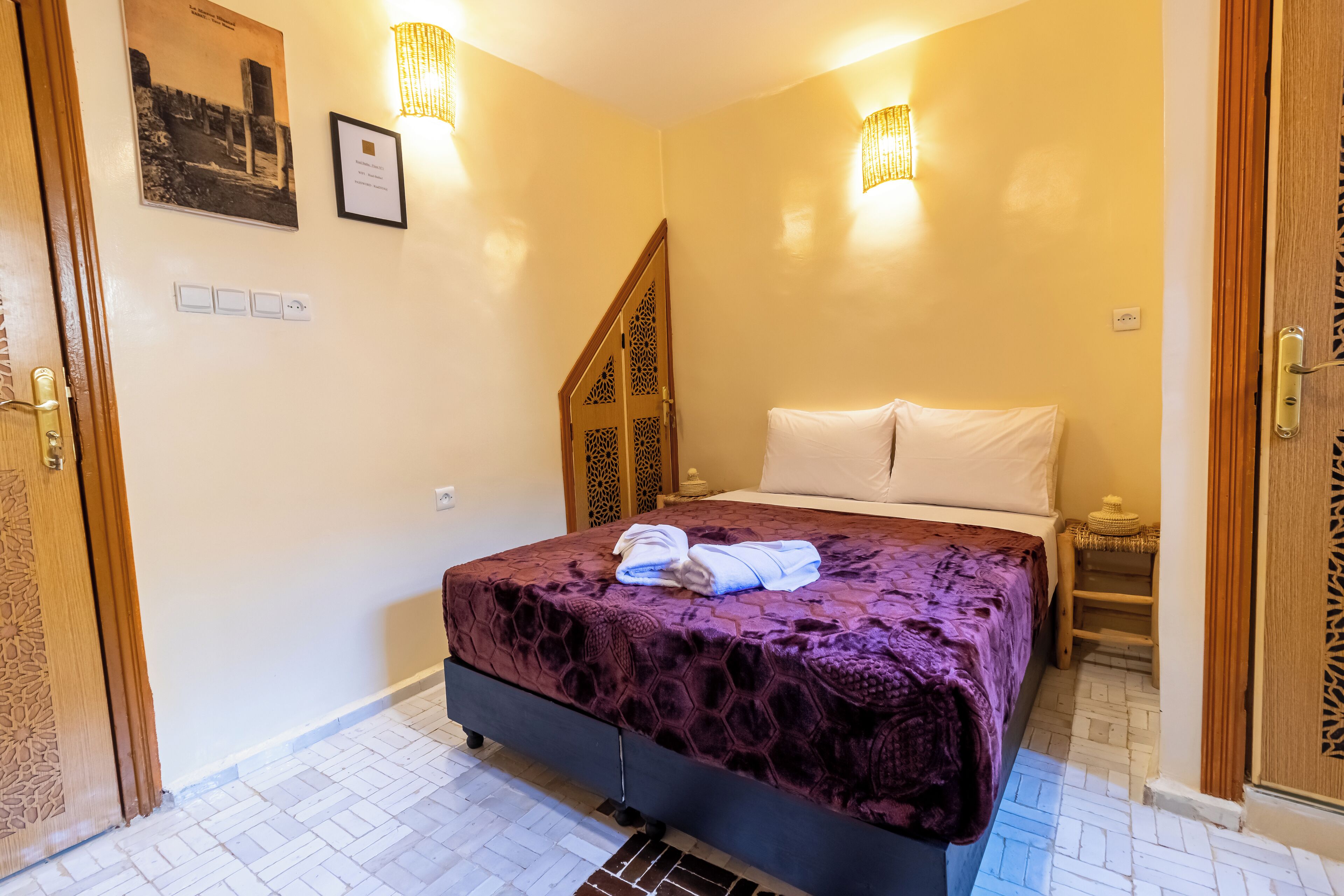 Une chambre à coucher comprenant un lit, une table de chevet, un luminaire fixé au mur et une porte ornée de motifs complexes en bois.