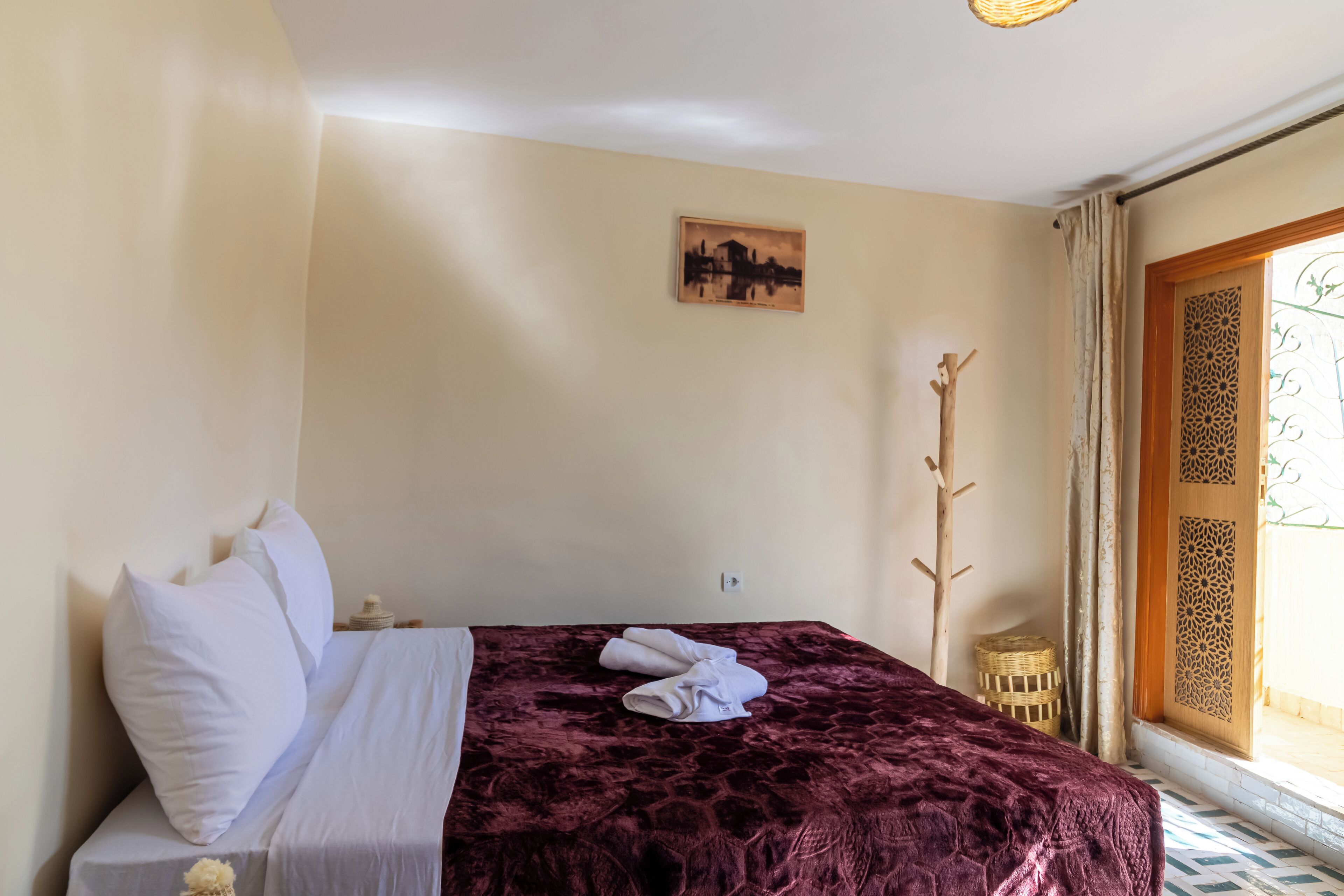 A neatly made bed with white linens and a dark red blanket, a small bedside table with a lamp, a wall-mounted picture, and a view through a door with decorative panels.