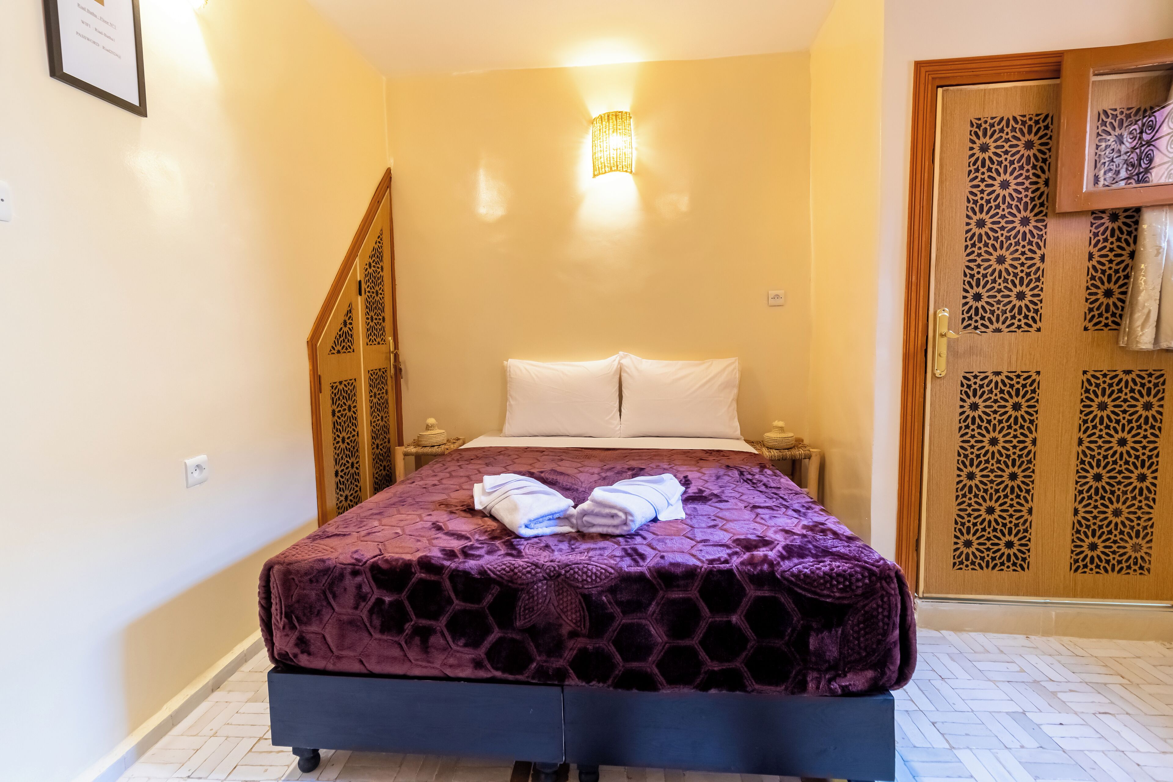 Quarto com cama, mesa de cabeceira, luminária na parede, porta com detalhes ornamentais e escada.