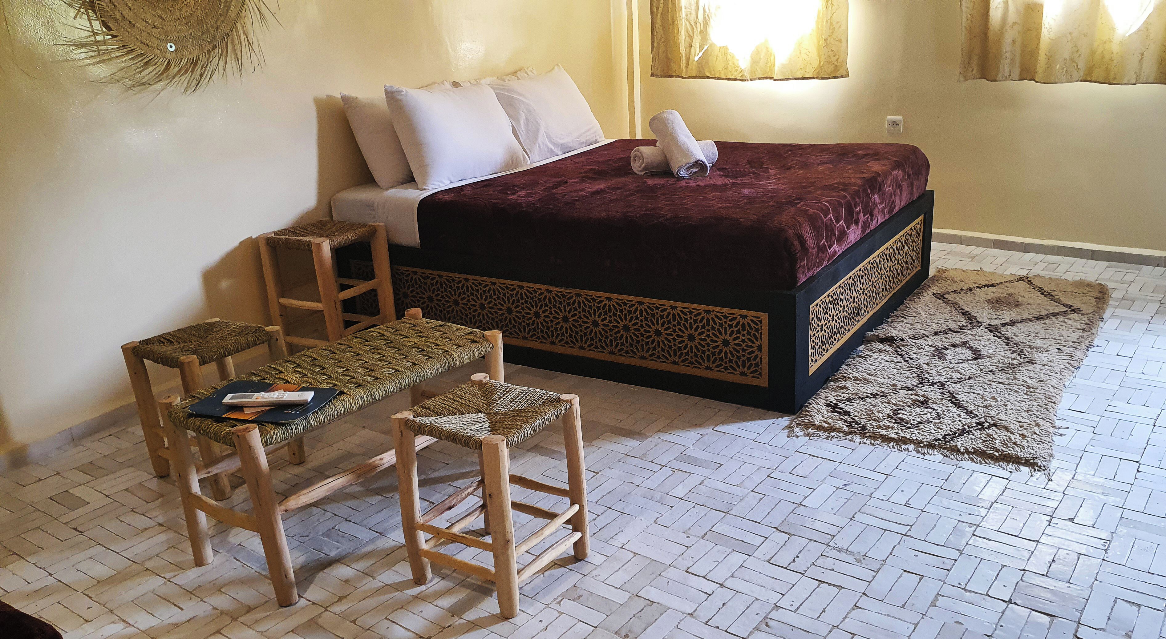 Un dormitorio con una cama, dos sillas de madera, una mesita pequeña, un tapete estampado y un panel decorativo en la pared.