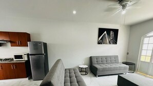 Una sala moderna con un sofá gris, una mesa de centro redonda y un reposapiés negro. Se ve un área de cocina con un microondas y un refrigerador al fondo.