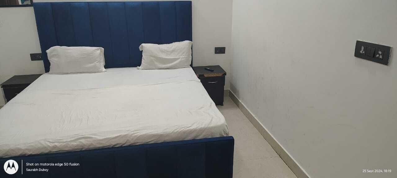 Una cama bien tendida con sábanas blancas y un cabecero azul en una habitación de hotel. Hay dos mesitas de noche, una con una lámpara y la otra con un enchufe.