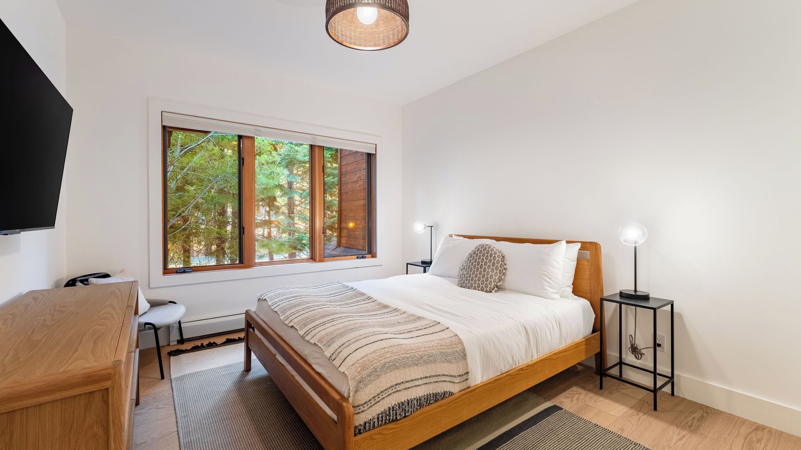 A bedroom with a large window, a bed, bedside tables, a desk, and a TV.