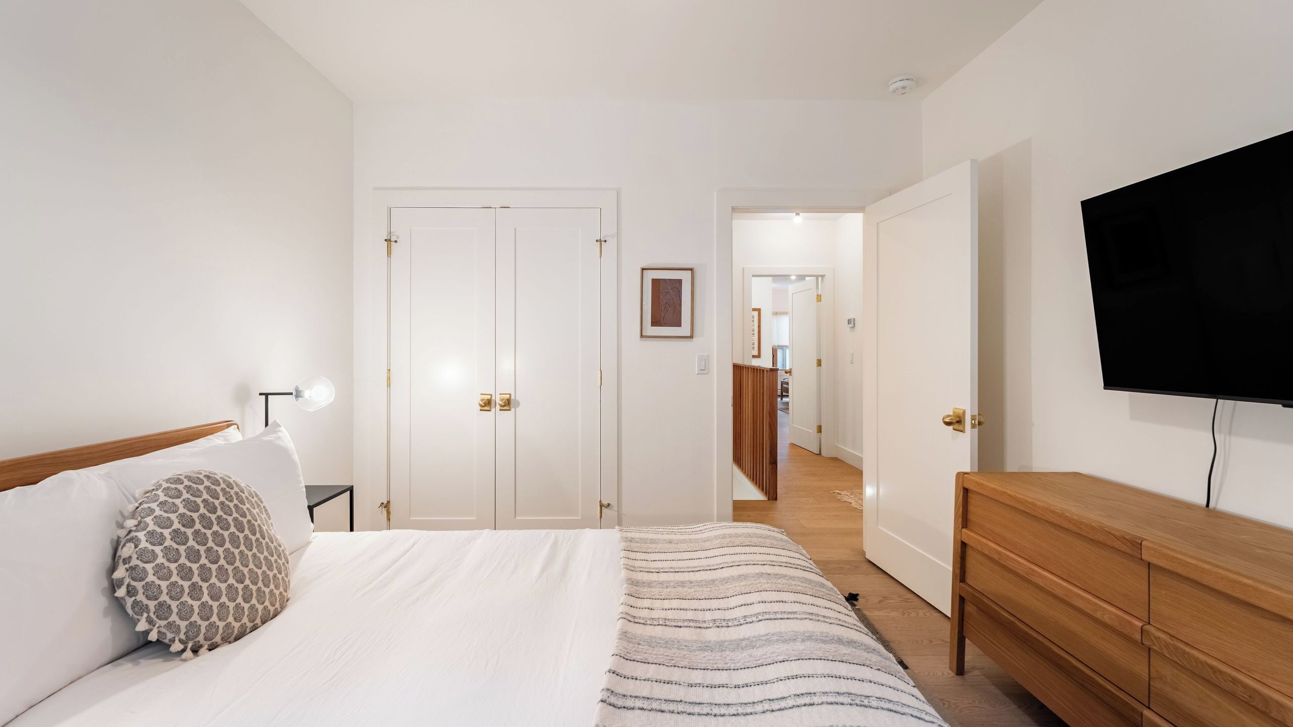 A bedroom with a bed, a dresser, a television, and a hallway leading to a kitchen.