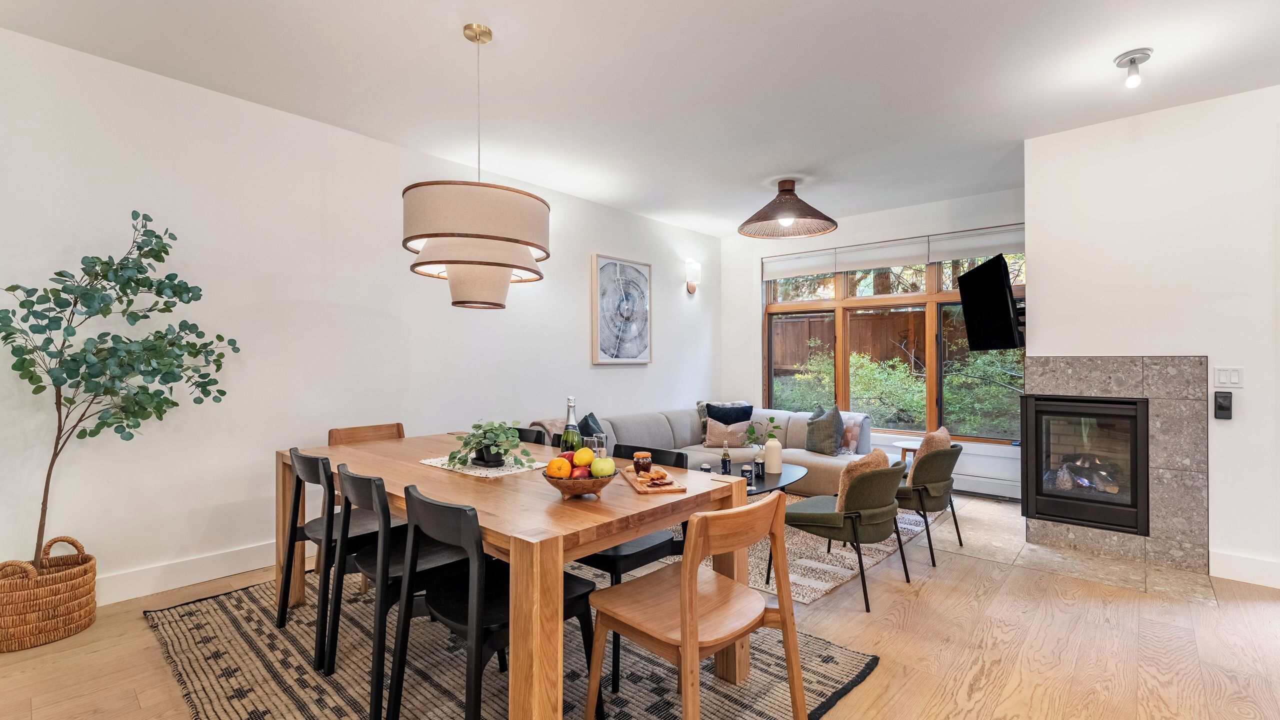 A modern dining area with a wooden table, black chairs, and a fruit bowl. There is a sofa and chairs in the background, a fireplace, and large windows with a view of greenery.