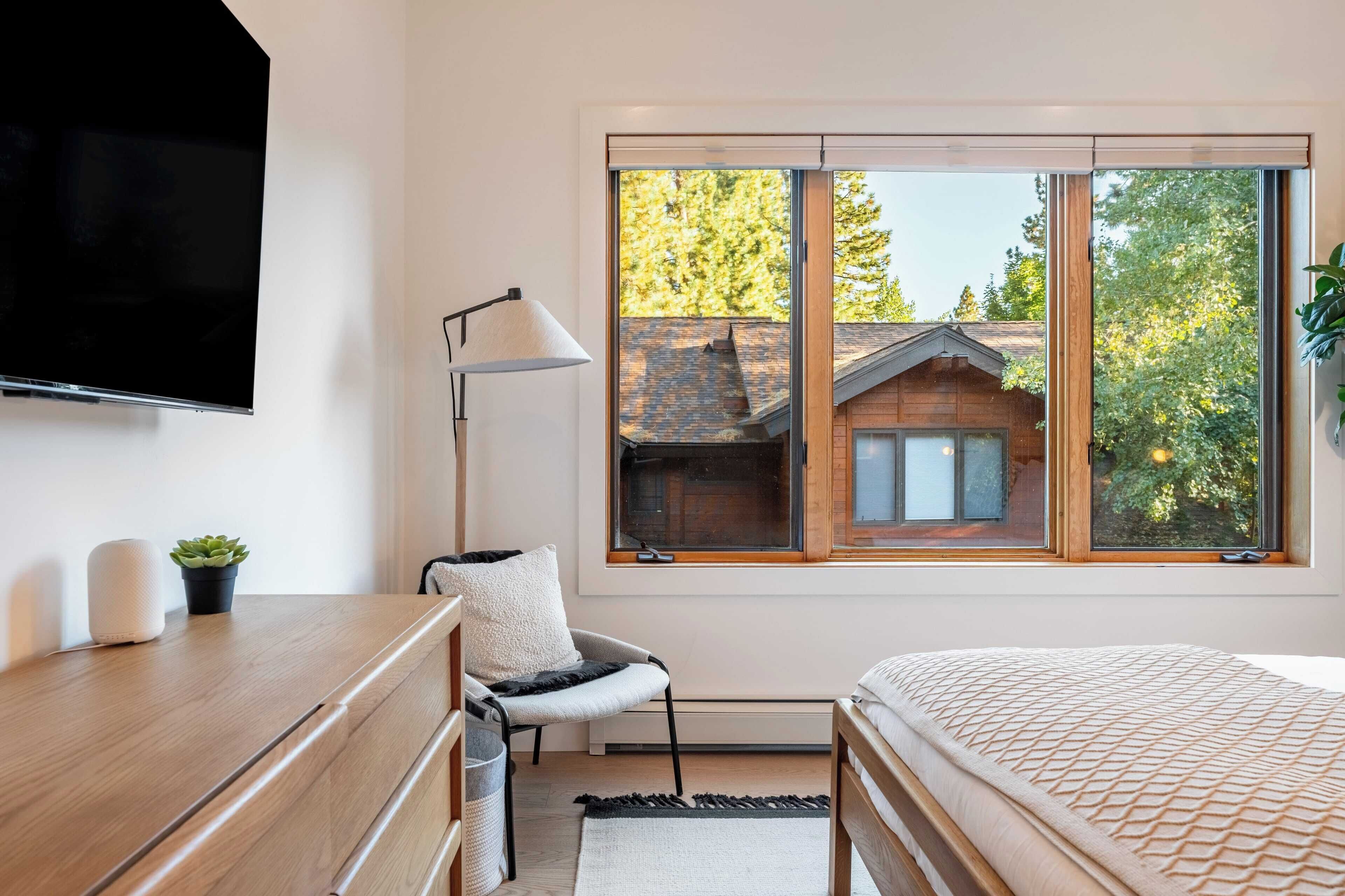 A bedroom with a large window, a bed, a chair, a desk, a TV, and a plant.