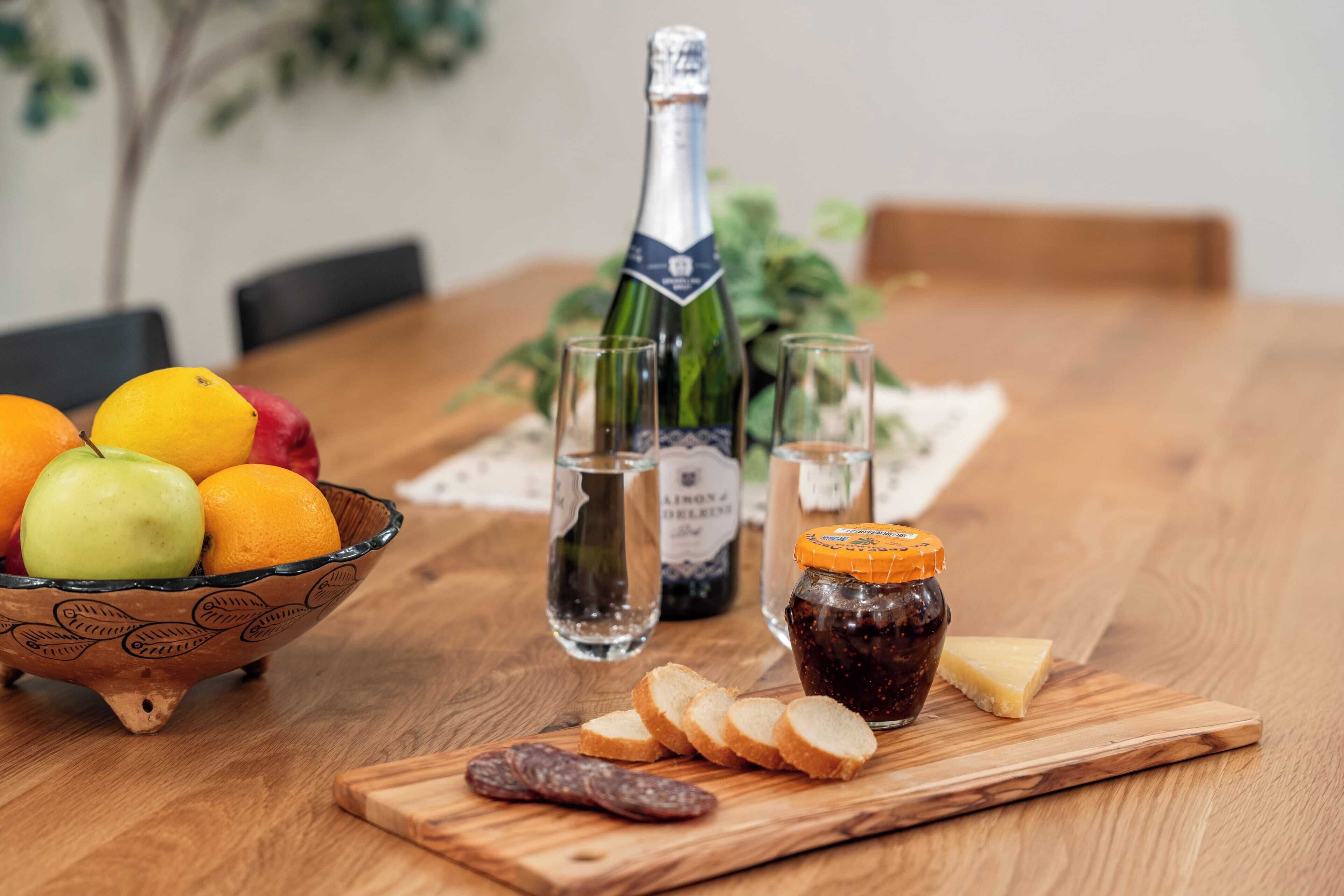A wooden table with a bowl of fruit, a bottle of champagne, glasses, and a jar of jam.