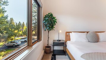 Une chambre avec une grande fenêtre, une table de chevet, une lampe, un lit avec une literie blanche et une plante en pot.