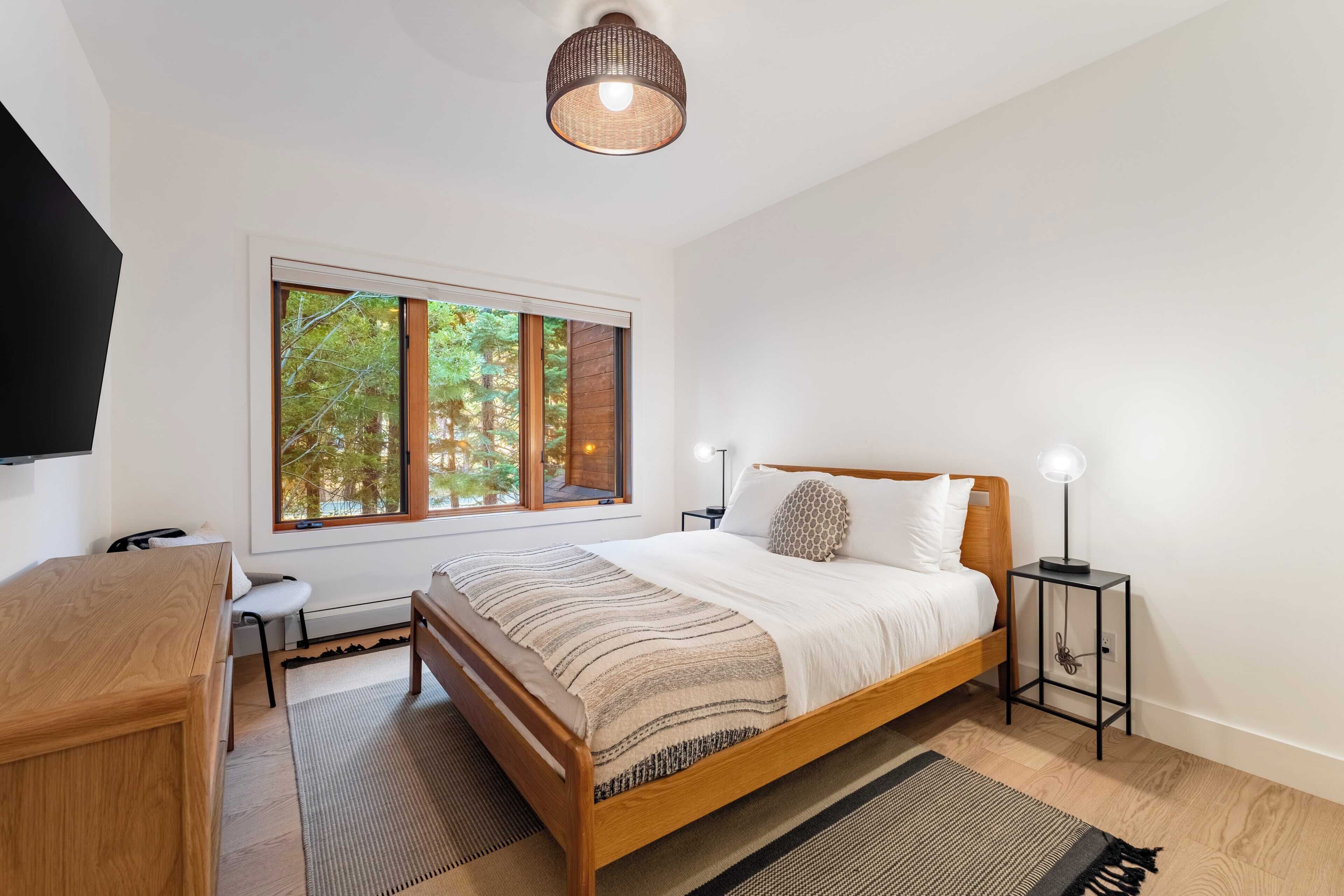 A bedroom with a large window, a bed with a wooden frame, a bedside table with a lamp, and a desk with a chair.