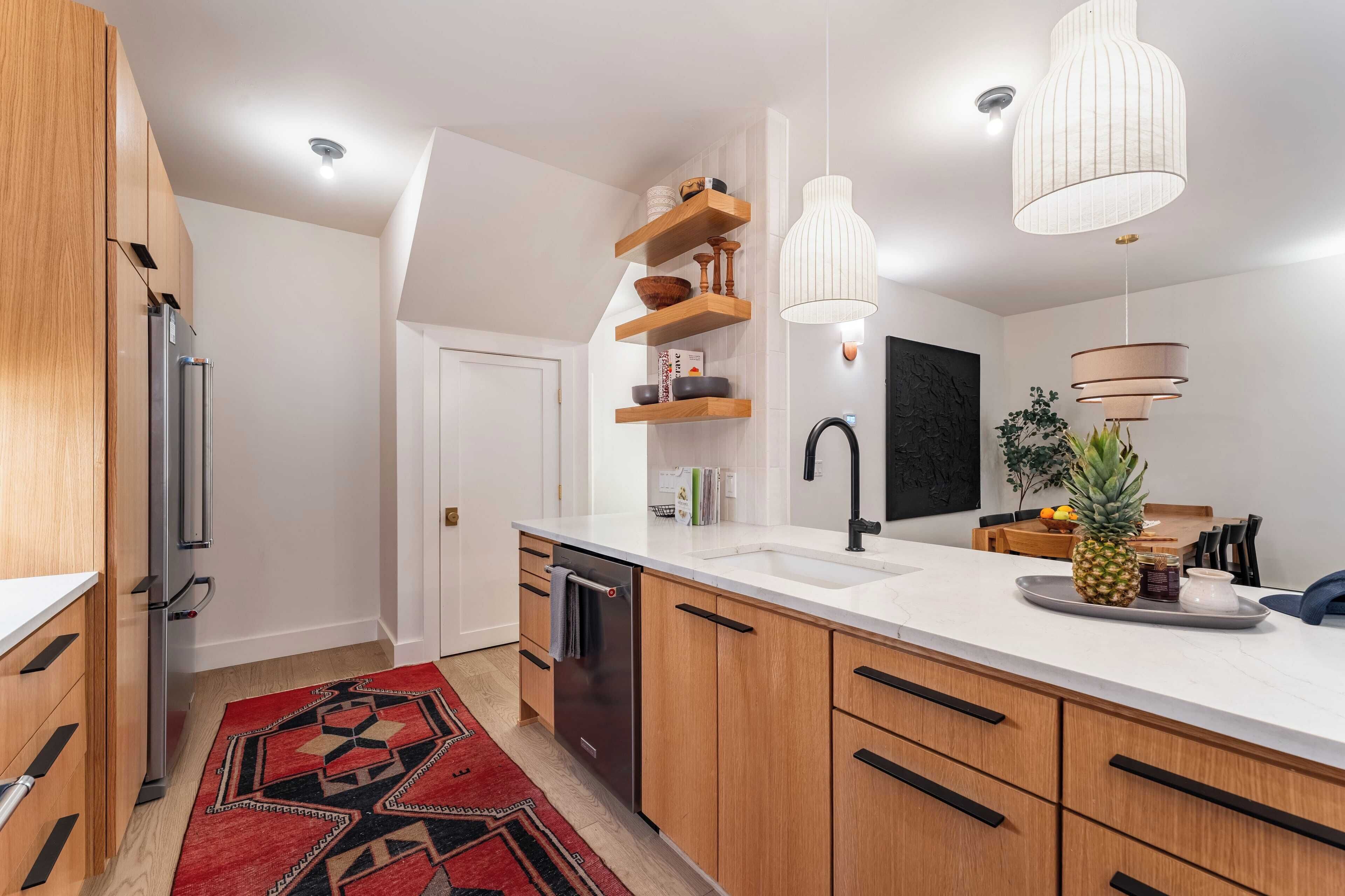 Eine moderne Küche mit Holzausziehtüren, einem Edelstahl-Kühlschrank, einem Geschirrspüler und einer weißen Arbeitsplatte. Es gibt Pendelleuchten, eine schwarze Spritzschutzverkleidung und einen roten Teppich mit Muster.