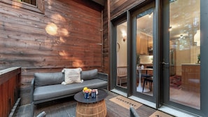 Une terrasse en bois avec un canapé, une table ronde avec des boissons, et une porte-fenêtre coulissante donnant sur un espace cuisine.