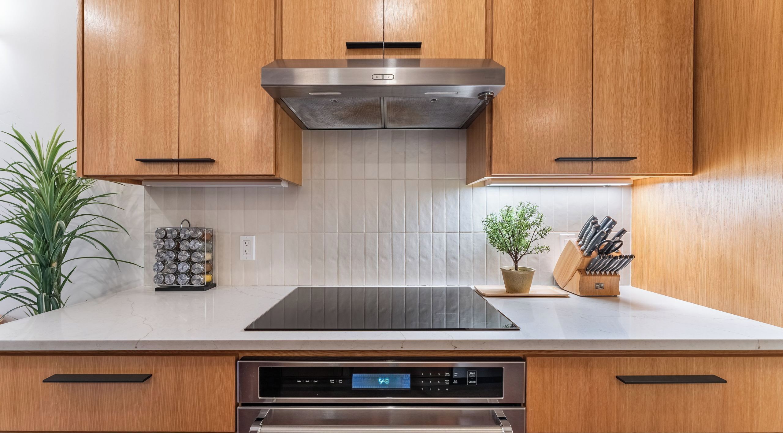 A kitchen with wooden cabinets, a stainless steel range hood, a digital oven control panel, a knife block, and a potted plant.