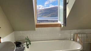 A bathroom with a bathtub, a window with a view of hills and water, and a plant on the counter. - Highland Rooms - Room Only No Breakfast (Ullapool)