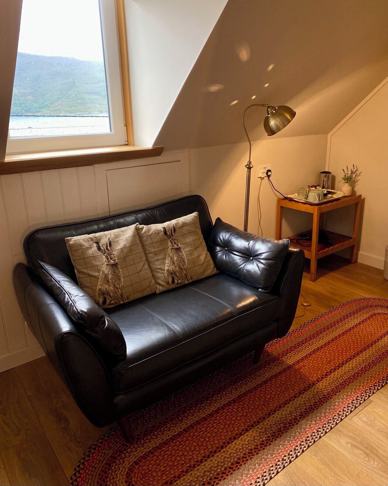 Un canapé en cuir noir agrémenté de coussins à motifs, une table d’appoint en bois avec un plateau, une lampe sur pied et une vue sur l’extérieur par une fenêtre.