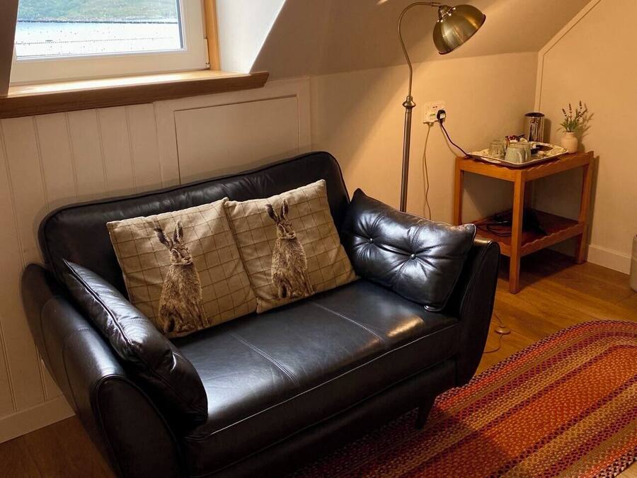 Un canapé en cuir noir agrémenté de coussins à motifs, une table d’appoint en bois avec un plateau, une lampe sur pied et une vue sur l’extérieur par une fenêtre.