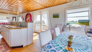 Ein geräumiges Zimmer mit Holzboden, einem großen Bett mit gemustertem Überwurf, einem Essbereich mit weißen Stühlen und einer Küche mit Spüle und Schränken.