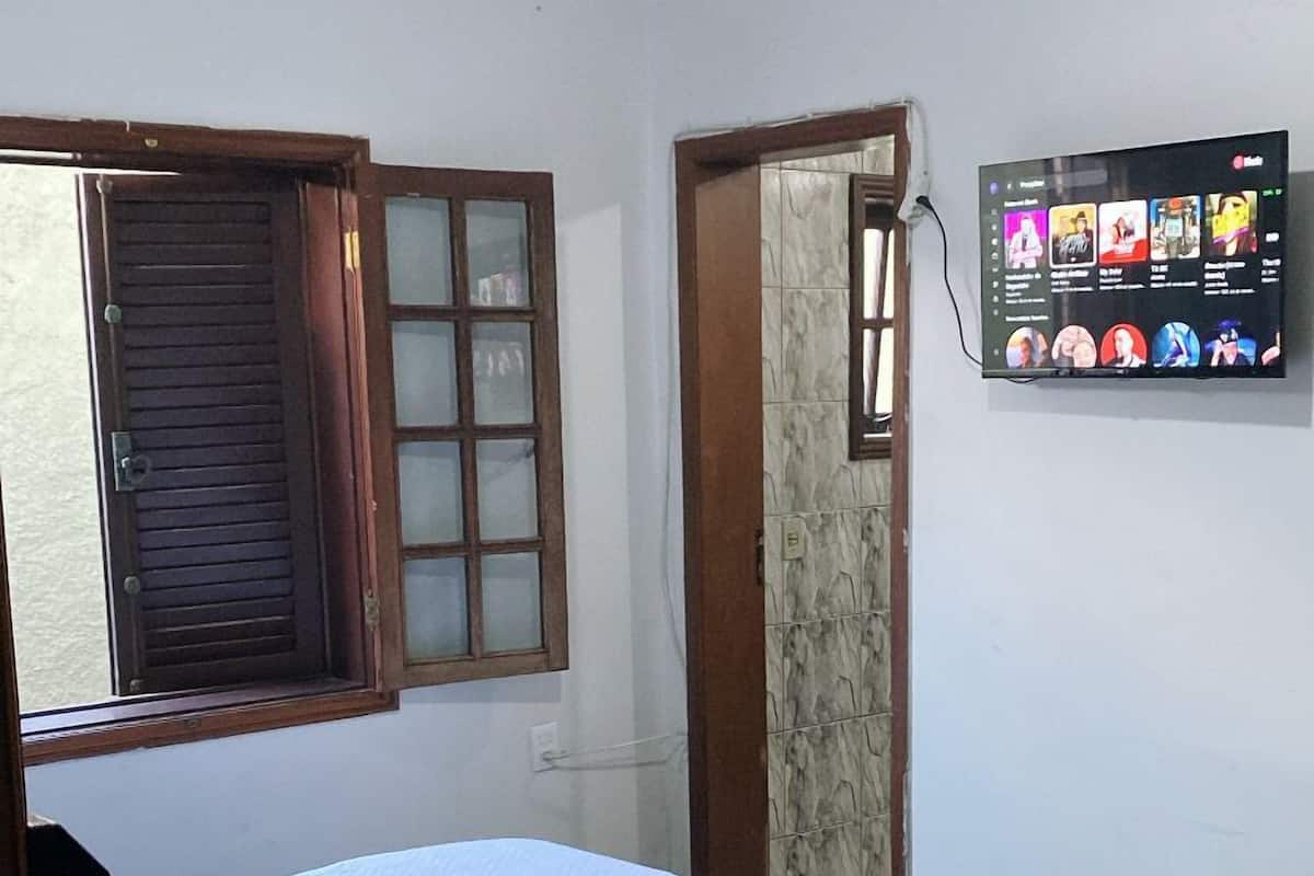 A bedroom with a bed, a window with wooden shutters, a wall-mounted TV, and a ceiling fan.