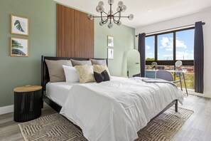 A modern bedroom with a large bed, a wooden sideboard, a round side table, and a view of the outdoors through a large window.