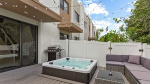 A modern outdoor deck with a hot tub, wicker furniture, and string lights.