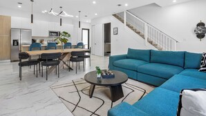 A modern living room with a teal sofa, a dining area with blue chairs, a kitchen with white cabinets, and a staircase.