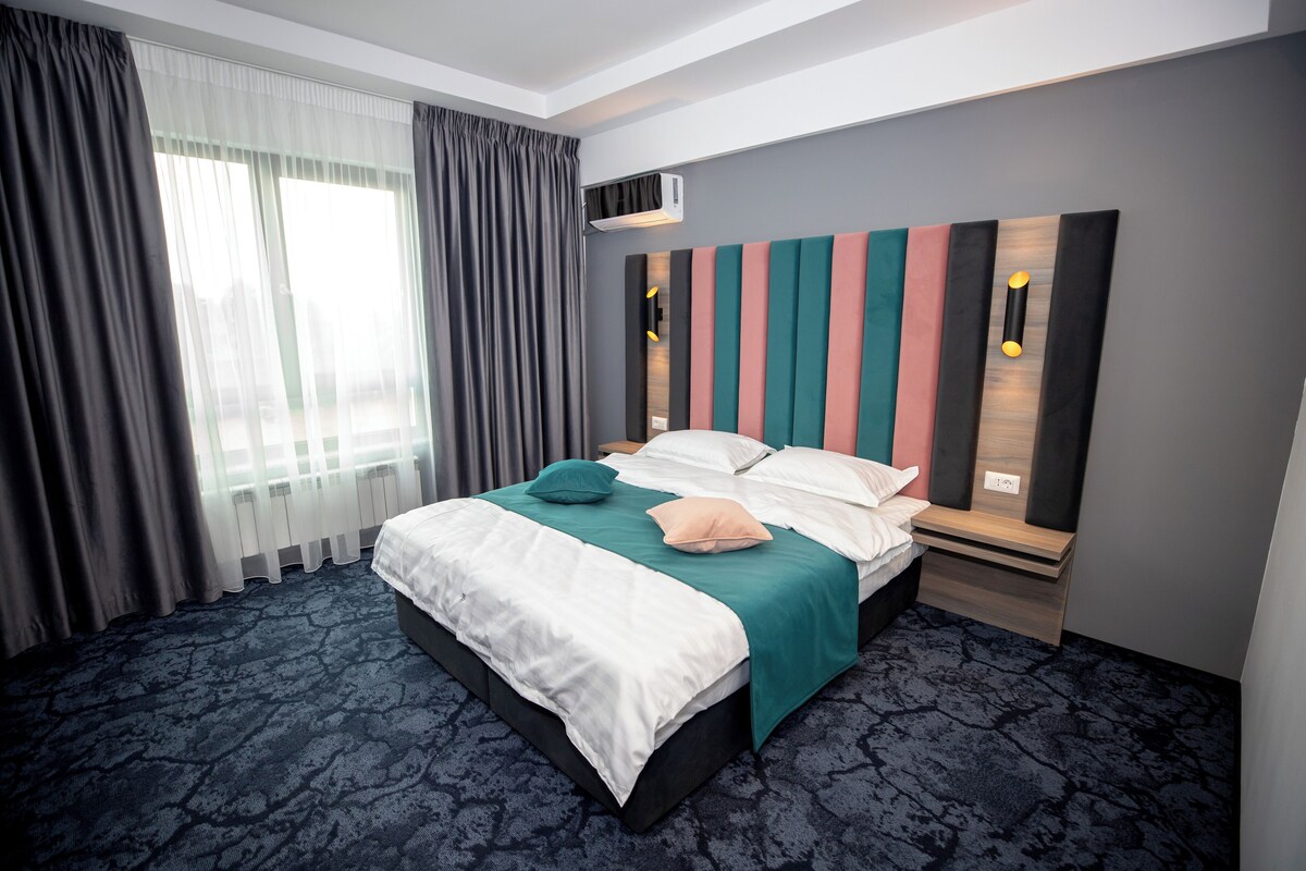 A modern hotel room with a large bed, a headboard with vertical stripes, a window with curtains, and a wall-mounted air conditioner.