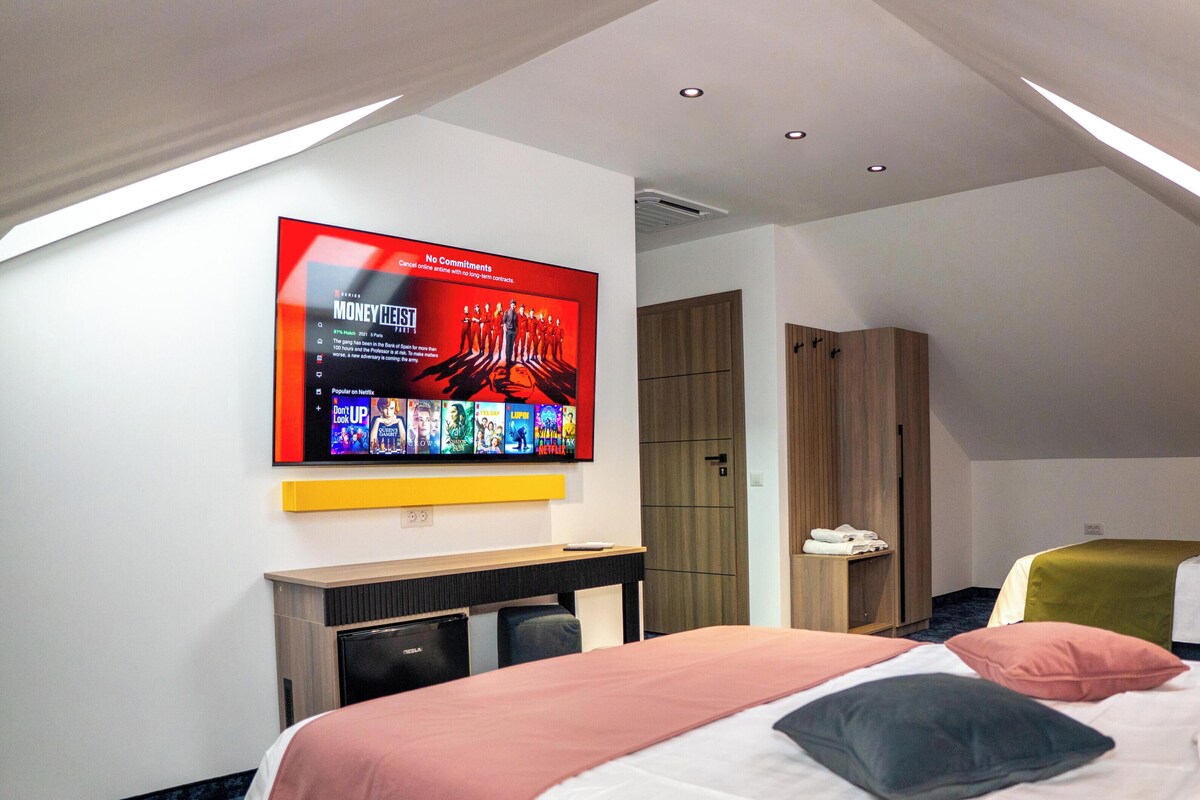 A modern hotel room with a large TV displaying a movie, a Tesla charging station, and a loft-style ceiling.