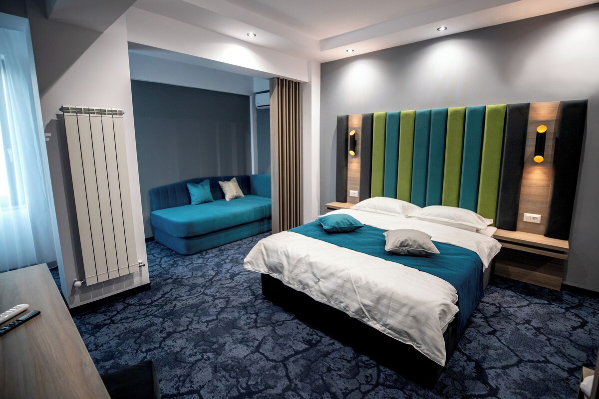 A hotel room with a large bed, a blue sofa, a wooden nightstand, and a wall-mounted light fixture.