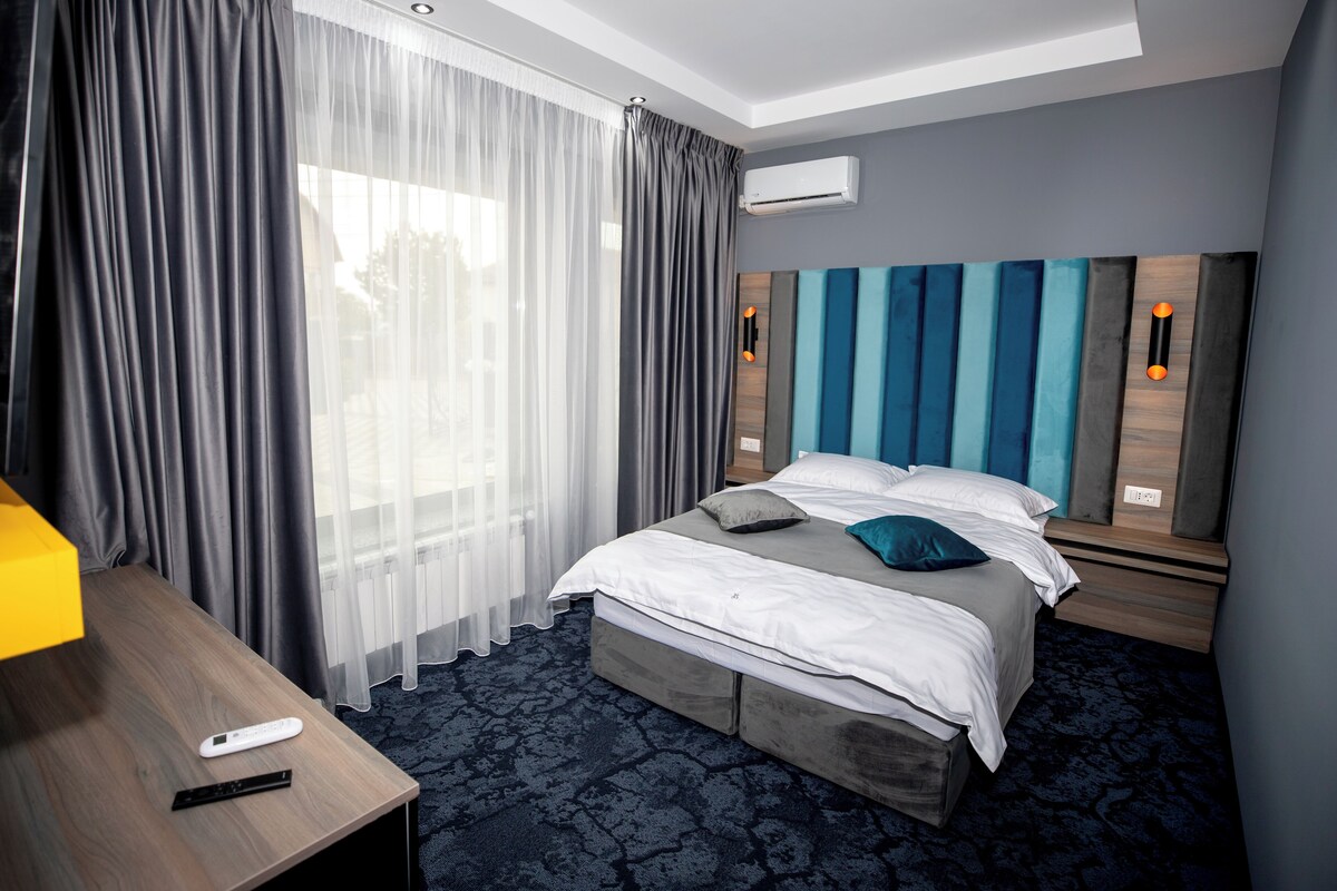 A modern hotel room with a large bed, a wooden nightstand, a desk with a lamp, and a window with sheer curtains.