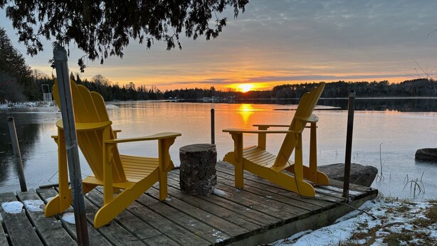 Deux chaises Adirondack jaunes sont placées sur une jetée en bois, offrant une vue sur un coucher de soleil au-dessus d’un lac.