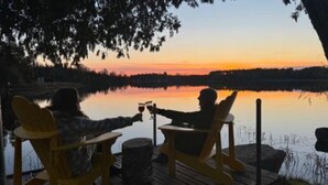 Deux personnes trinquent avec des verres de vin à une table au bord du lac, assises sur des chaises, et contemplent un lac paisible et un coucher de soleil.