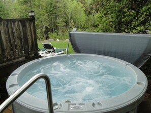 Un bain à remous doté d’un escalier en métal, entouré d’une terrasse en bois et d’une zone herbeuse avec des chaises longues.