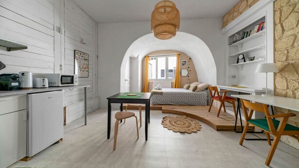 Een compacte, moderne keuken met witte kastjes, een zithoek met een ronde tafel en stoelen, en een gezellig slaapkamer met een bed en een groot raam.