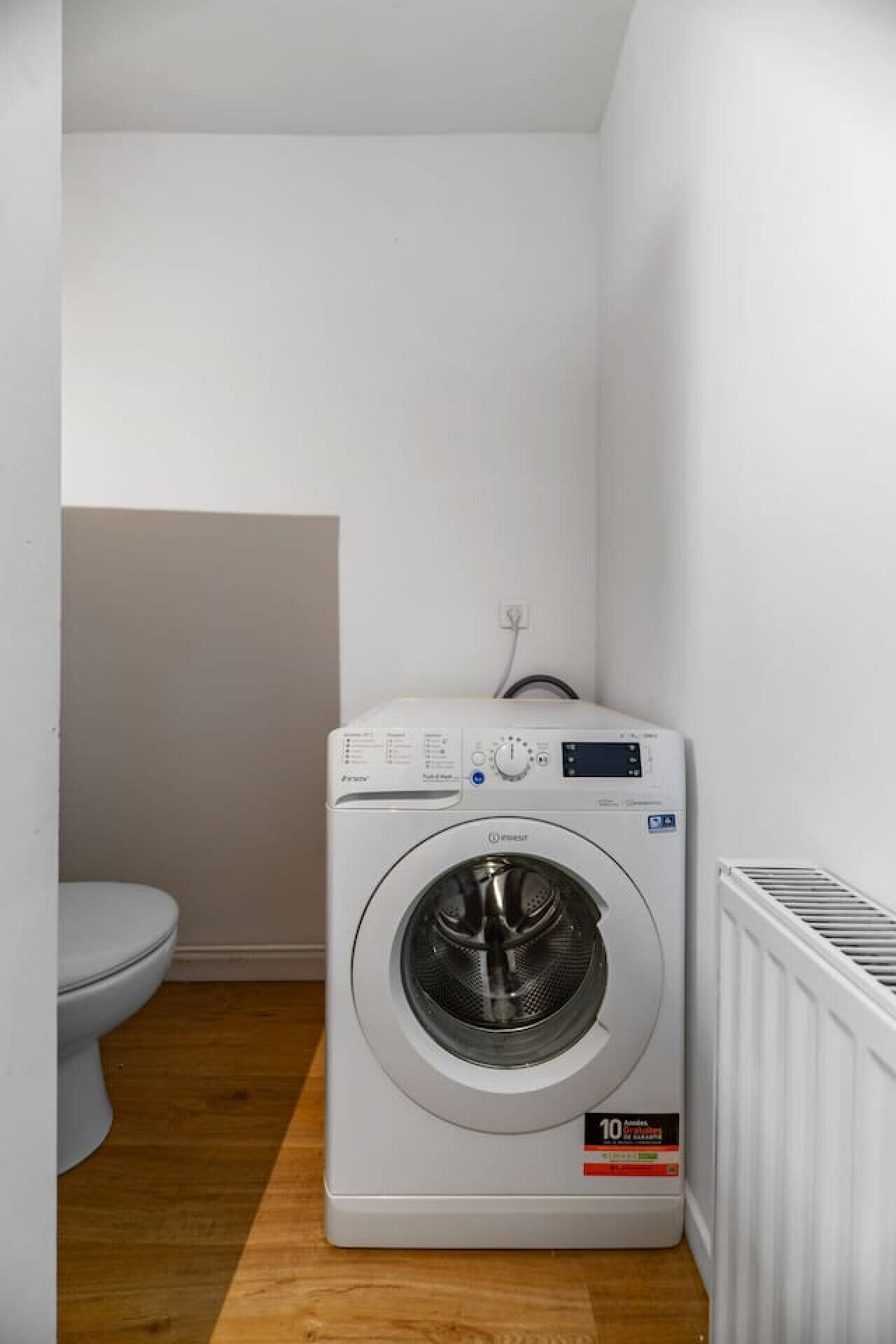 Een compacte, witte wasmachine in een kleine wasruimte met een houten vloer en een wit toilet.