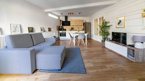 Een moderne woonkamer met een grijze hoekbank, een eethoek met een witte tafel en stoelen, en een keuken met houten kastjes en een zwarte kookplaat.