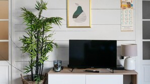 Een woonkamer met een televisie op een witte kast, een grote groene plant en een ingelijst foto aan de muur.