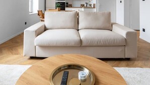 A modern living room with a beige sofa, a wooden coffee table with a remote and a container, and a kitchen in the background. - Air-cond and designer apartment (Vichy)