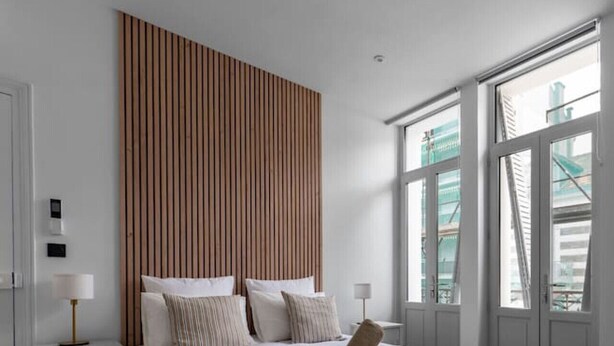 A modern bedroom with a large bed, wooden panel headboard, bedside tables with lamps, and large windows with a view of buildings. - Air-cond and designer apartment (Vichy)