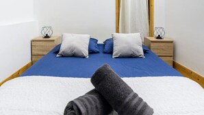 A neatly made bed with blue bedding, two pillows, and rolled towels. There are two bedside tables with decorative items. A window with a curtain is visible in the background. - Le Cosy - 11 Rue de la Boucherie - Clermont (Clermont-Ferrand)