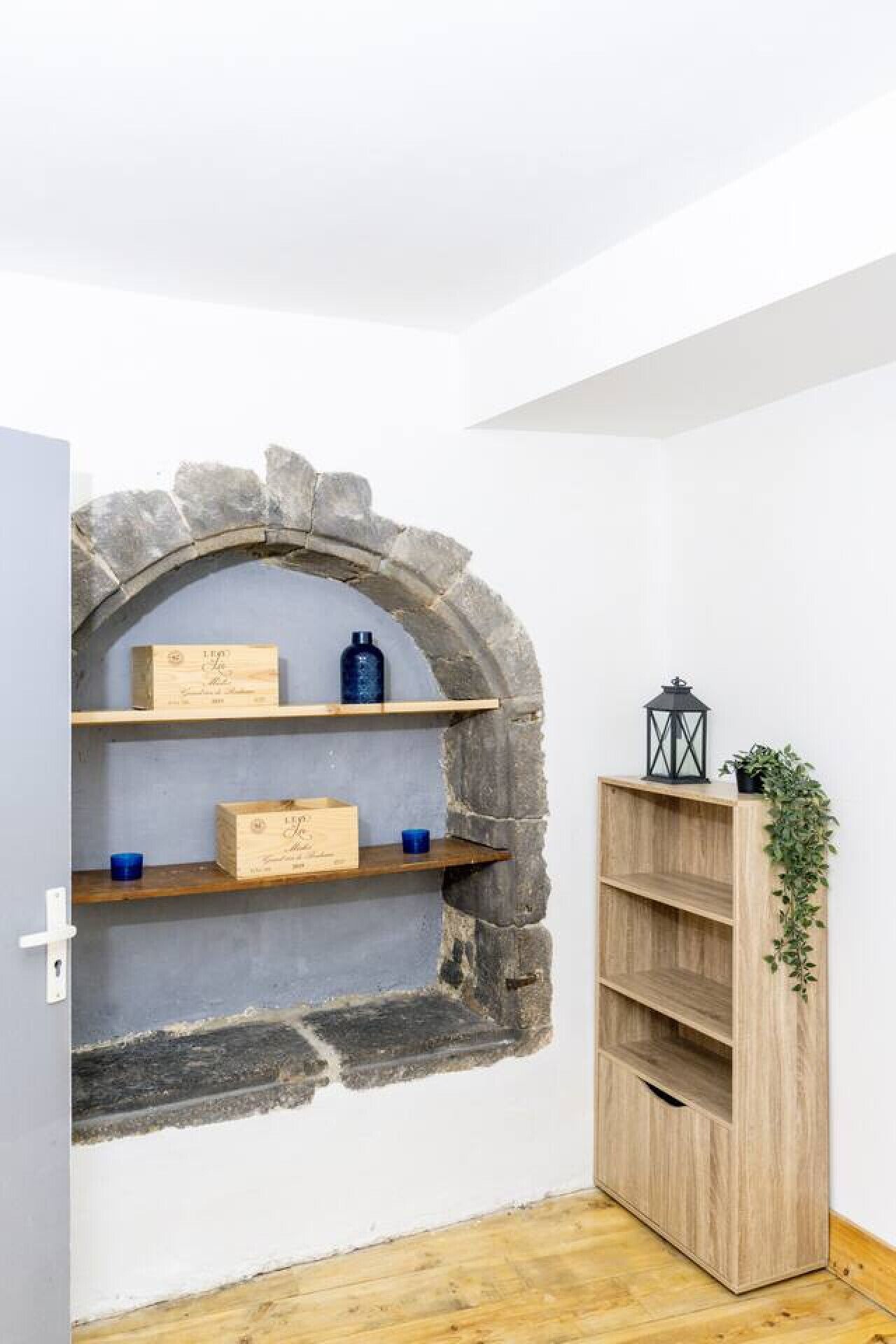 Een kamer met een stenen boog die leidt naar een plankenwand met houten dozen en blauwe bakken. Ook zichtbaar is een houten boekenkast en een lantaarn.