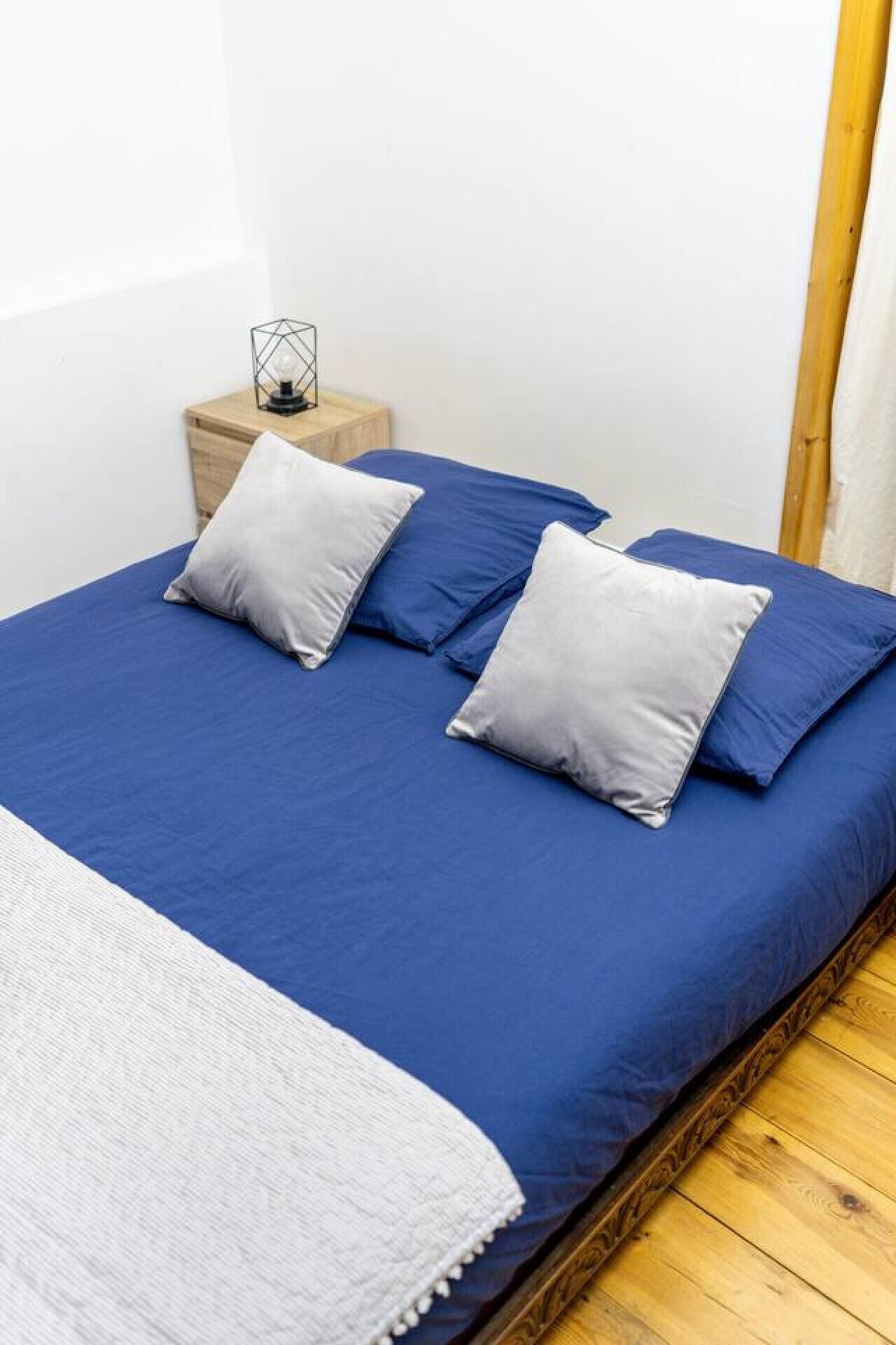 Una cama bien hecha con ropa de cama azul y dos almohadas, una mesita de noche con lámpara y suelo de madera.