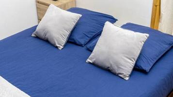 Una cama bien hecha con ropa de cama azul y dos almohadas, una mesita de noche con lámpara y suelo de madera.