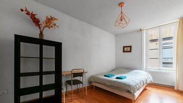 A bedroom with a bed, a desk, a chair, a window with blinds, and a hanging light fixture.
