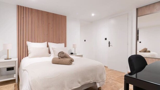 Ein modernes Hotelzimmer mit einem großen Bett, einer Wandpaneele aus Holz, einem kleinen Tisch mit Stuhl und einem durch eine Glastür einsehbaren Badezimmer.