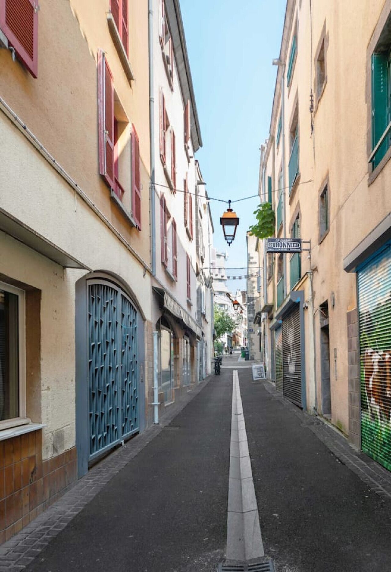 Un carrer estret flanquejat per edificis, alguns amb persianes i tendals, i un fanal a la part superior.