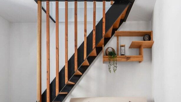 Un dormitorio tipo loft moderno con escalera de madera, cama blanca con almohadones estampados y una pequeña estantería con objetos decorativos.