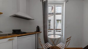 Una cocina compacta con armarios blancos, una campana extractora de acero inoxidable, una pequeña mesa de comedor con dos sillas y vistas a la calle a través de una ventana.
