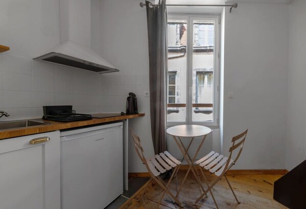 Una cocina compacta con armarios blancos, una campana extractora de acero inoxidable, una pequeña mesa de comedor con dos sillas y vistas a la calle a través de una ventana.