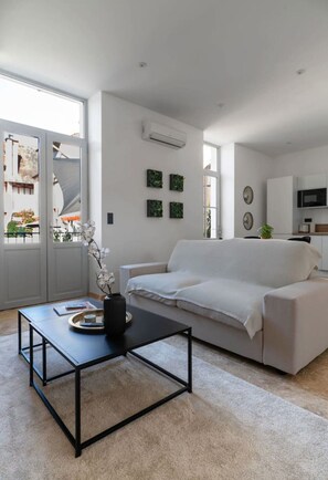 A modern living room with a white sofa, a black coffee table, and a small plant. There is a door leading to a balcony with a view of buildings.