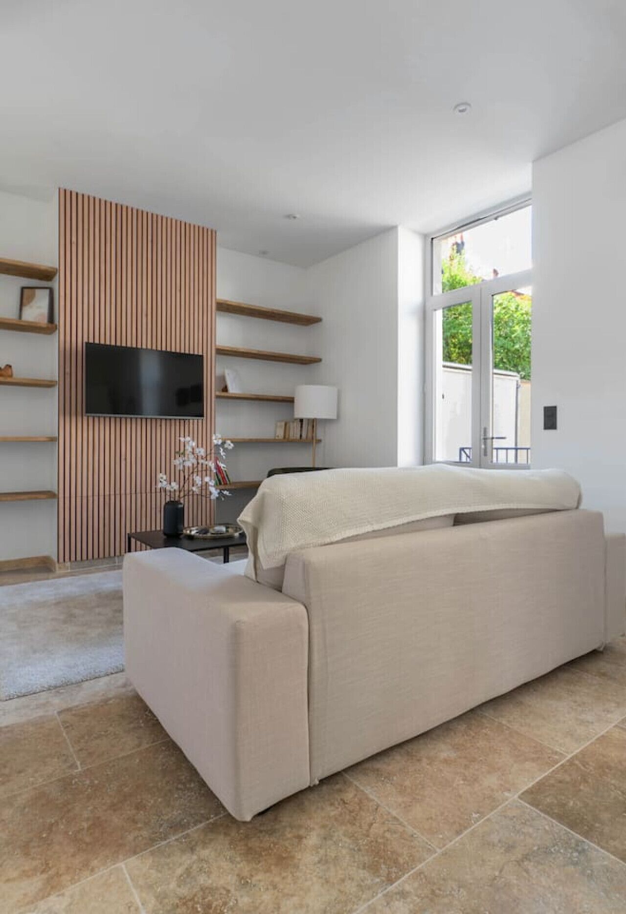 A modern living room with a sofa, a flat-screen TV mounted on a wooden panel, and a large window providing a view of the outdoors.