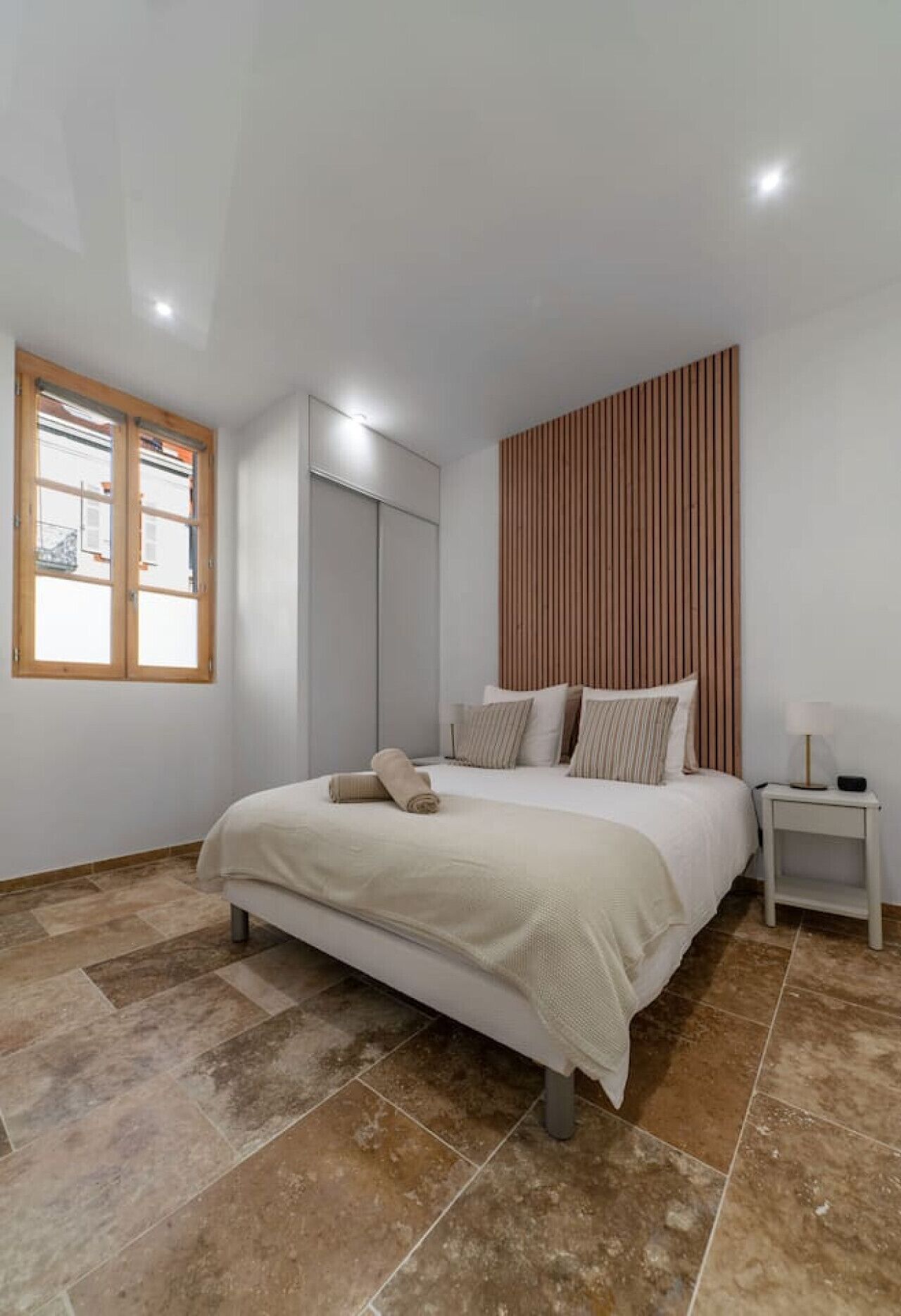 A bedroom with a bed, a nightstand, a wooden slat wall panel, a window with wooden frames, and a tiled floor.
